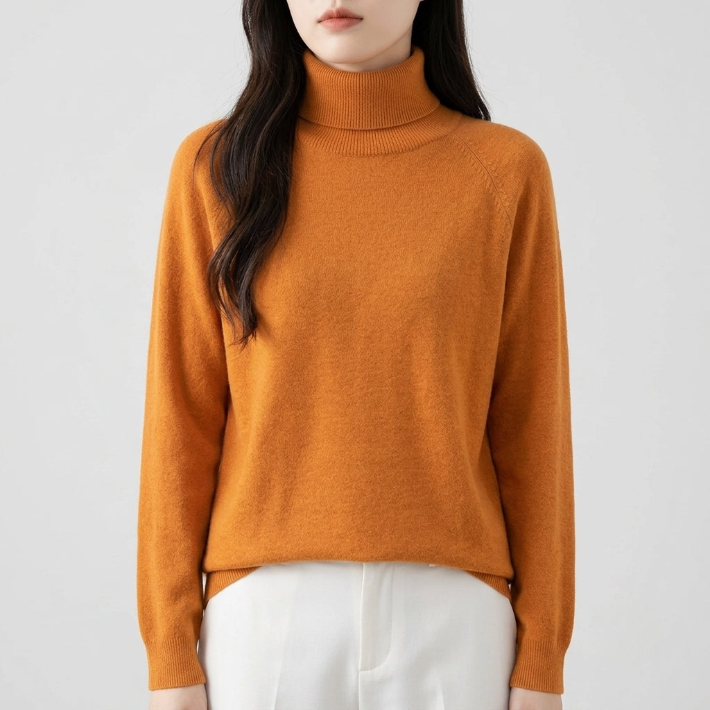 A woman wearing a cropped, long-sleeve turtleneck sweater in a warm camel orange color, paired with white pants, against a plain light gray background.