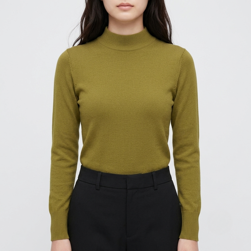 A woman wearing a fitted long-sleeve mock neck knit sweater in olive green, paired with black trousers, against a light gray studio background.