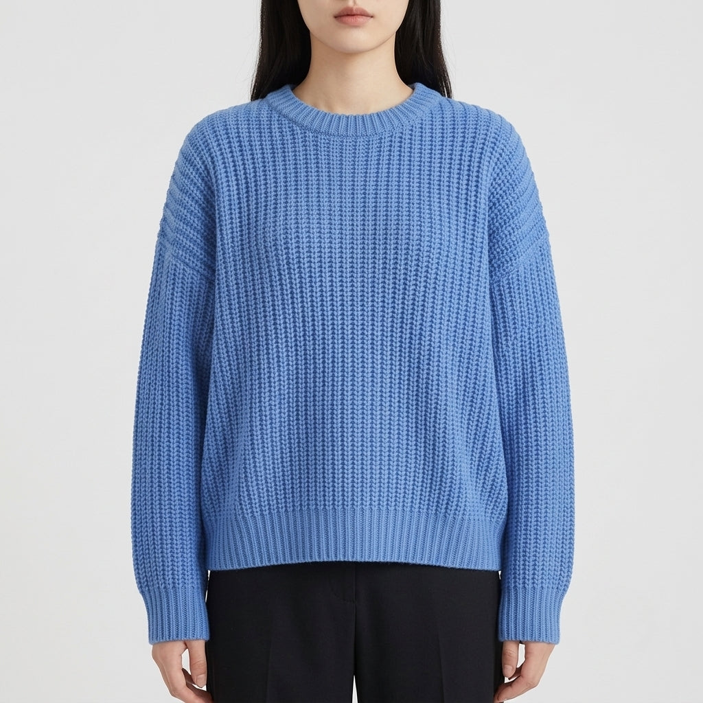 A woman wearing a light blue ribbed knit sweater with black pants, shown from the neck down against a plain light gray studio background.