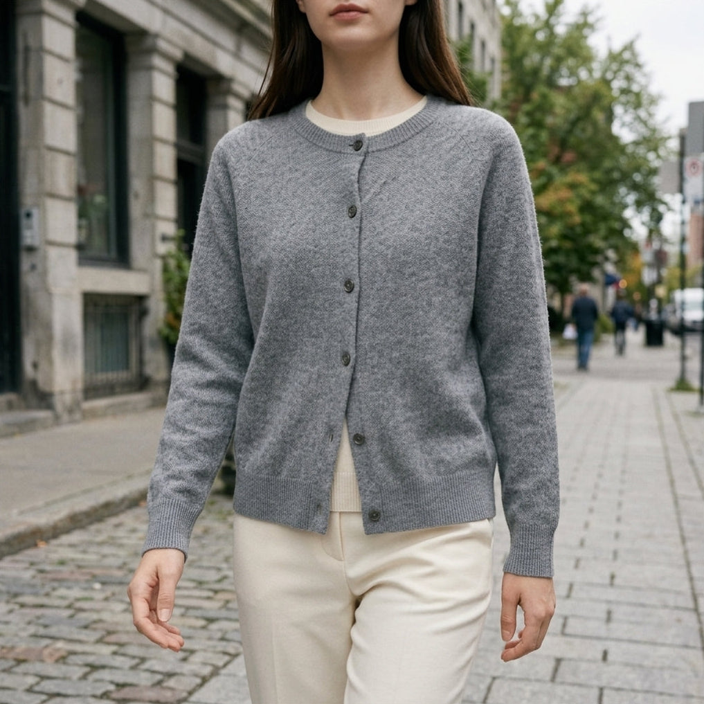 A woman wearing a gray button-front cardigan over a cream top and cream pants, standing on a cobblestone city street.